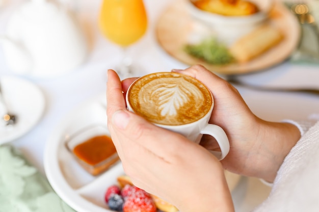 Женские руки, держа чашку свежесваренного кофе с красивым листом латте-арт из пены на фоне стола с тарелкой еды. концепция утреннего завтрака и позднего завтрака