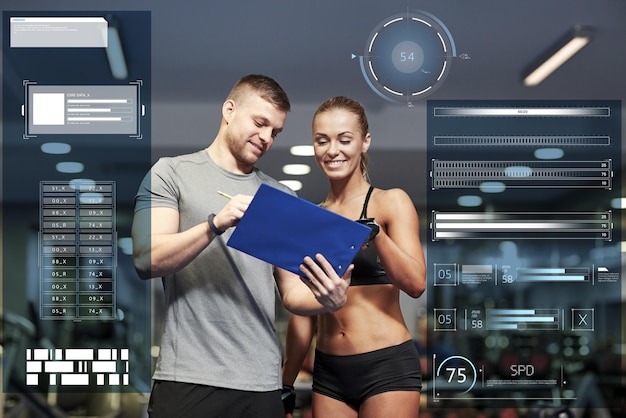 Photo fitness, sport, exercising and people concept - smiling young woman and personal trainer with clipboard writing exercise plan in gym over virtual charts