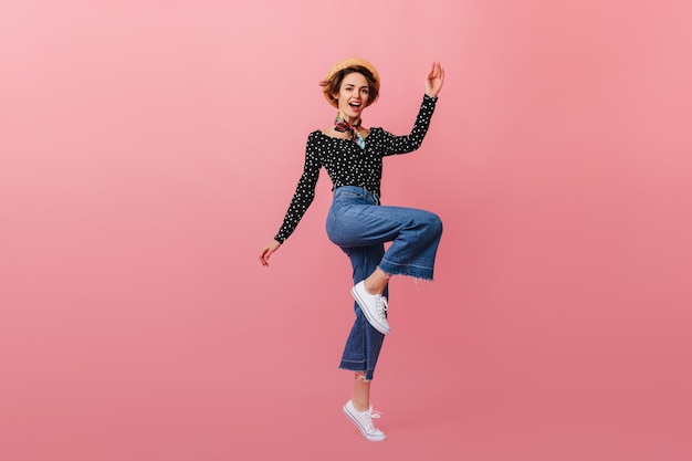 Full length view of jumping woman in straw hat Studio shot of girl in vintage jeans dancing on pink background