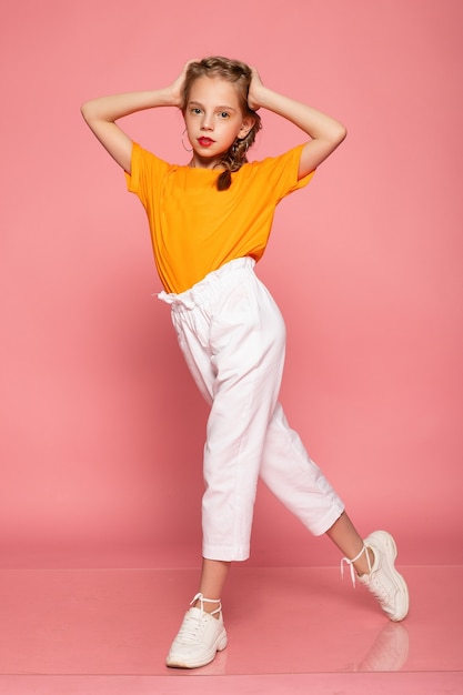 Full length little girl on pink studio wall. wearing an orange T-shirt and white pants and white sneakers.  