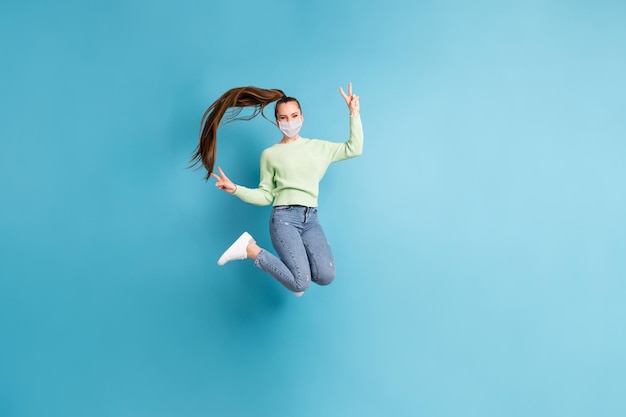 Полная длина фото прыгающей девушки с длинными волосами, показывающая v-знак носить медицинскую маску, изолированную на ярко-синем цветном фоне
