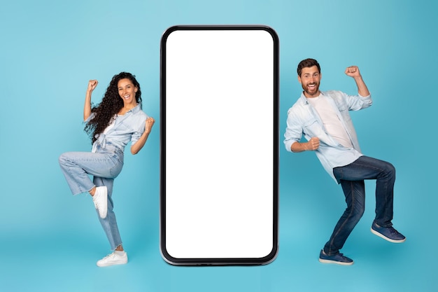 Gelukkig opgewonden jonge internationale man en vrouw dansen verheugt zich om te winnen in de buurt van grote smartphone met leeg scherm