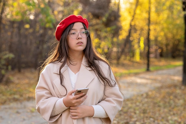 Девушка в очках, светлом пальто и красном берете идет в осенний парк и держит в руке телефон