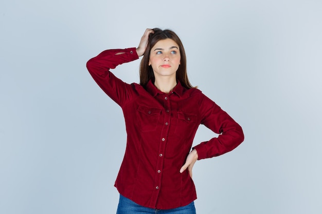 Photo girl holding hand on head, looking away in burgundy shirt and looking thoughtful.