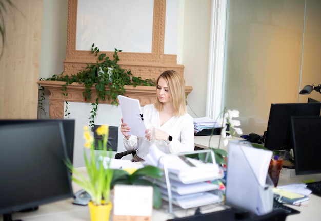 Фото Красивая женщина смотрит на экран компьютера, держа в руках лист бумаги в офисе