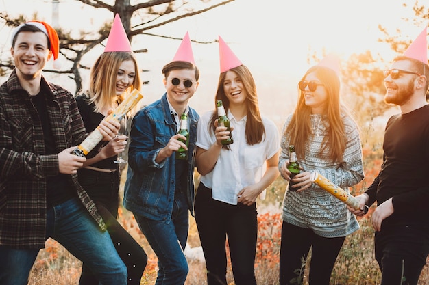 Группа молодых людей с бутылками пива и крекерами, смеющимися во время празднования Рождества на природе. Друзья с алкоголем и рождественскими крекерами