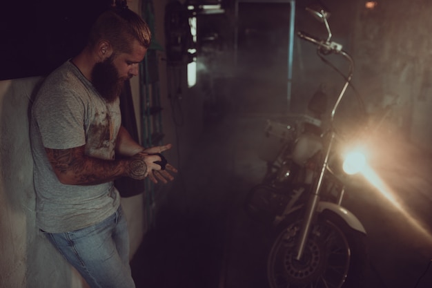 Красивый брутальный мужчина с бородой стоит в своем гараже на фоне мотоцикла и смотрит в сторону