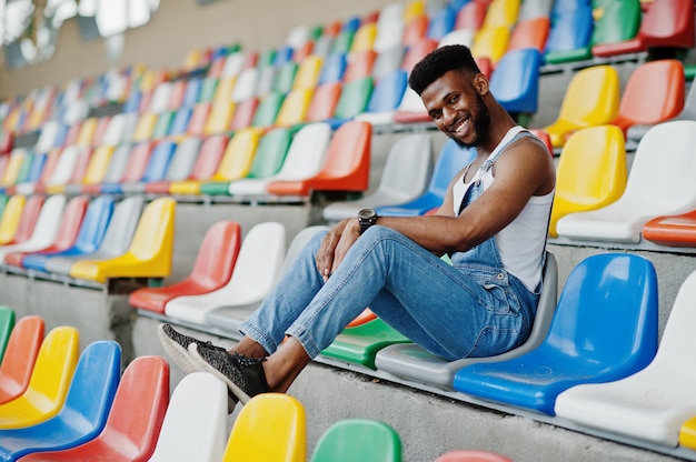 Красивый мужчина в джинсовых комбинезонах позирует на цветные стулья на стадионе Модный черный мужской портрет.