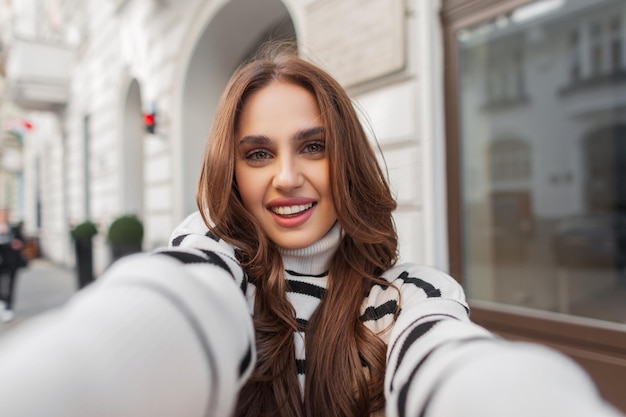 Фото Счастье молодая красивая женщина с милой улыбкой в модном полосатом свитере наслаждается путешествием и делает селфи на смартфоне возле белого старинного здания в европейском городе