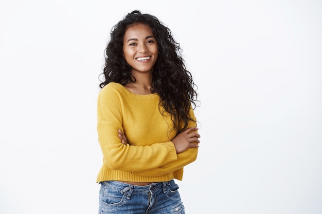 Счастливая привлекательная афро-американская женщина держит скрещенные руки и уверенно смеется, смотрит в камеру с восхитительной веселой улыбкой, носит желтый свитер на белой стене, концепция благополучия
