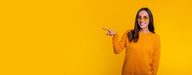 Счастливая великолепная молодая брюнетка в свитере и солнцезащитных очках указывает в сторону, пока она позирует изолированной на желтом фоне