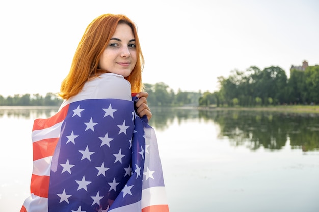 Photo happy smiling red haired girl with usa national flag on her shoulders. positive young woman celebrating united states independence day.