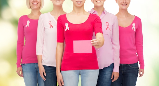 Photo healthcare, people and medicine concept - close up of smiling women in shirts with pink breast cancer awareness ribbons and blank paper card over green background