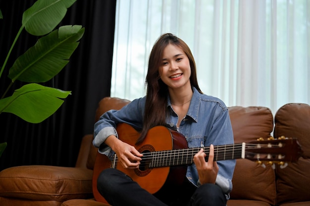 그녀의 현대 빈티지 거실에서 기타를 연주하는 진 재킷을 입은 힙스터 아시아 여성