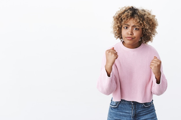 I will show you, come closer. Portrait of angry and cute african american blonde in sweater frowning offended and sulking raising fists in boxing pose wanting punch and fight over white wall