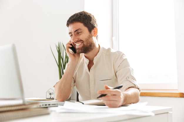 Изображение радостного бизнесмена 30-х годов в белой рубашке, говорящего по мобильному телефону, сидя за столом в современном офисе