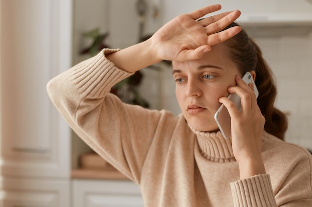 Фото Снимок в помещении: женщина в бежевом свитере сидит на кухне и разговаривает по смартфону, рассказывая о своей болезни, чувствует себя плохо, страдает от головной боли, держа руку на лбу