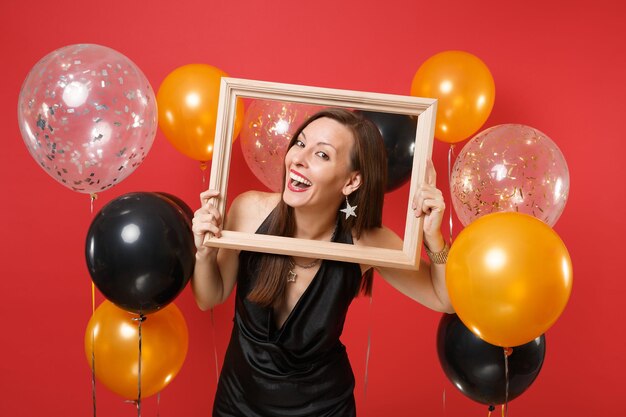 Радостная молодая женщина в маленьком черном платье празднует и держит рамку на ярко-красном фоне воздушных шаров. Международный женский день, с новым годом, концепция праздничной вечеринки макет дня рождения.