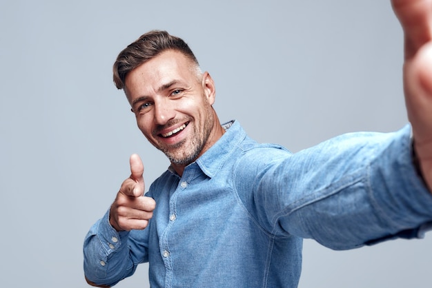 Photo keep smiling cheerful bearded man in shirt making selfie and gesturing while standing against grey