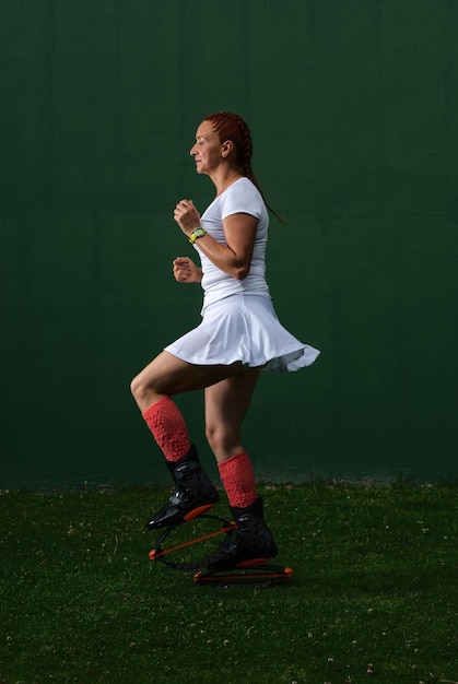 Латинская рыжая женщина в белой спортивной одежде делает тренировку в черных ботинках Kangoo Jumps на темно-зеленом фоне.