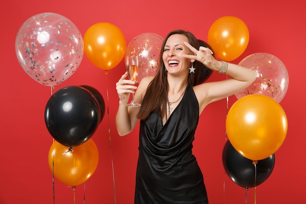 Смеющаяся девушка в черном платье празднует проведение бокала шампанского, показывая знак победы на ярко-красном фоне воздушных шаров. День святого Валентина, с новым годом, концепция праздничной вечеринки макет дня рождения.