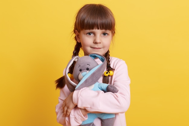 Фото Маленькая девочка обнимает своего мягкого кролика, очаровательная девочка с косичками, выражающая любовь своему любимому пушистому кролику у желтой стены, ребенок играет с игрушкой.