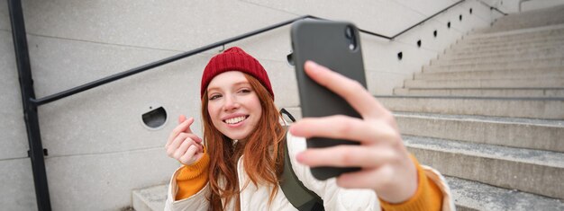 Фото Мобильный телефон и образ жизни людей. стильная рыжая девушка делает селфи на смартфоне, позирует для