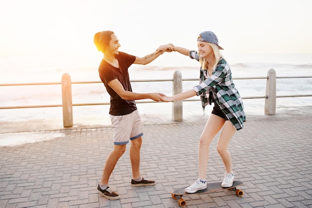 Теперь вы понимаете. Полнометражный снимок красивого молодого человека, который учит свою девушку кататься на скейтборде на набережной.