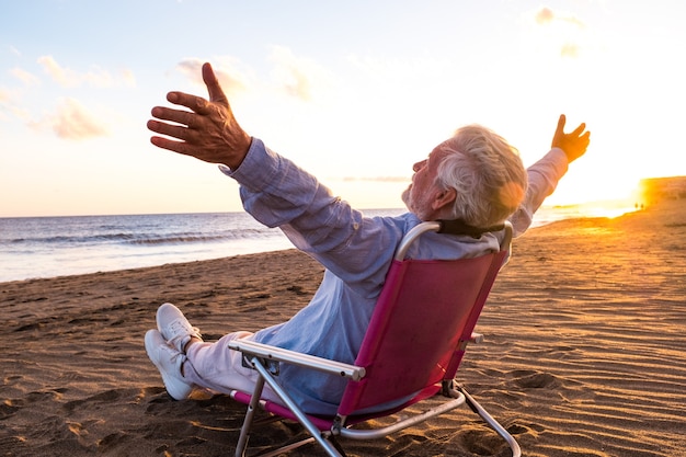 Photo one mature and old man enjoying the summer vacations alone at the beach sitting in a small chair looking to the sea. male person feeling free with opened arms. freedom concept.