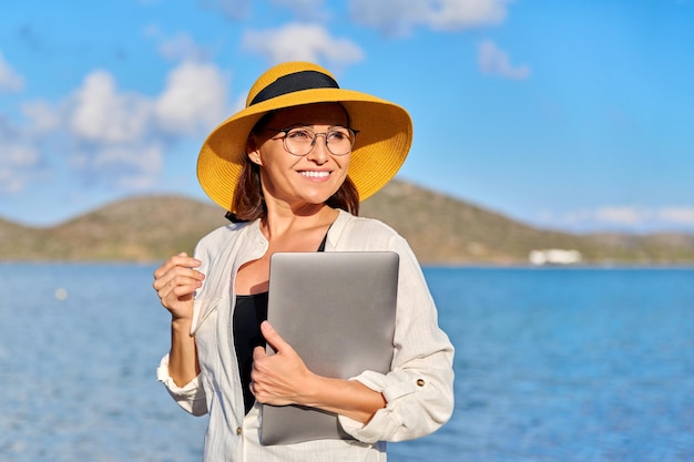 Открытый портрет зрелой женщины в шляпе на пляже с ноутбуком в руках