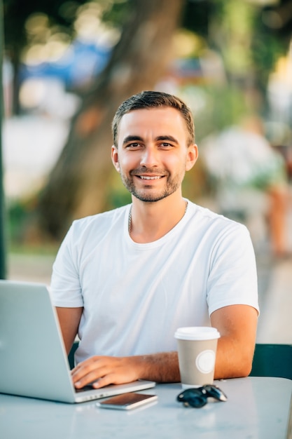 Photo outdoor portrait of young smiling european man sitting at cafe with his laptop