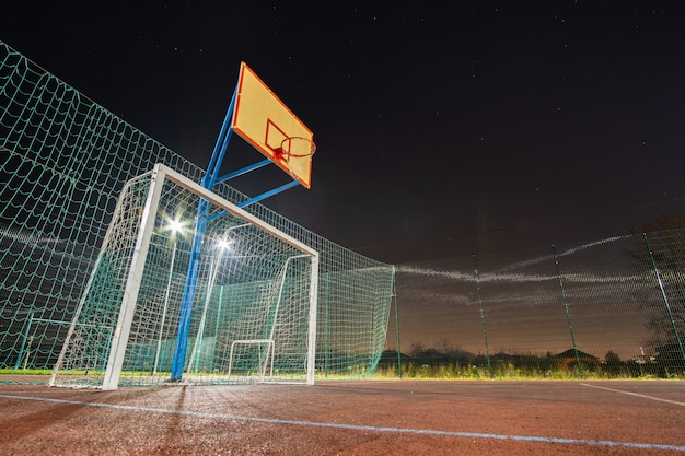 Photo outdoors mini football and basketball court with ball gate and basket surrounded with high protective fence brightly illuminated with spotlight lamps at night.