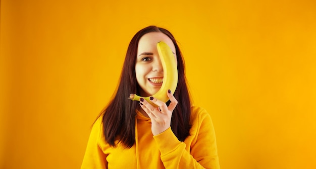 Портрет молодой женщины с бананом на желтом фоне Смешная женщина в желтой толстовке позирует с фруктами