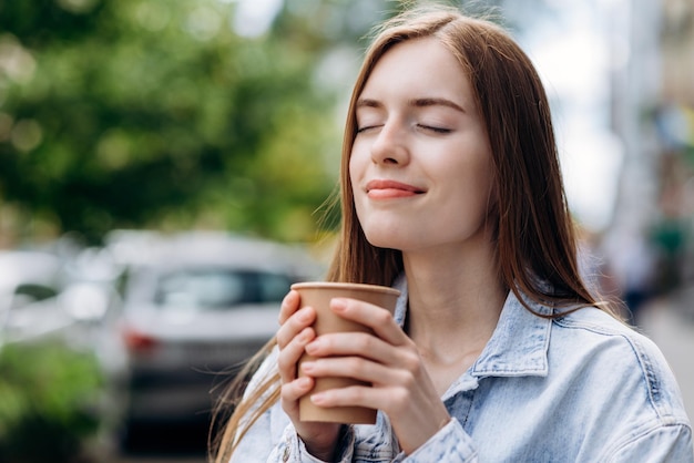 Portrait of young beautiful woman inhaling aroma and drinking coffee while walking around the city Model standing with closed eyes with coffee to go