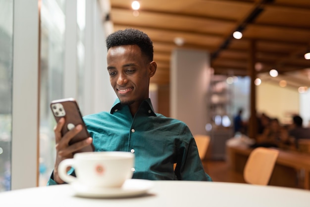 Портрет красивого молодого темнокожего мужчины в кафе с помощью телефона