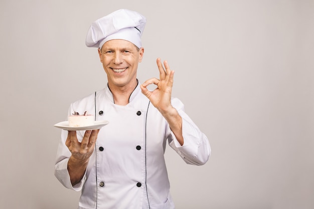 Портрет счастливого старшего мужчины шеф-повара, одетый в форму, держа тарелку с куском пирога