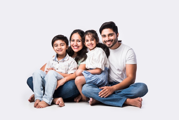 Photo portrait of indian asian young family of four sitting on white flour against white background, looking at camera