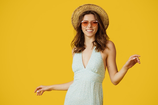 Фото Портрет красивой женщины в летнем платье соломенной шляпе и красочных солнцезащитных очках, позирующей и танцующей на желтом фоне