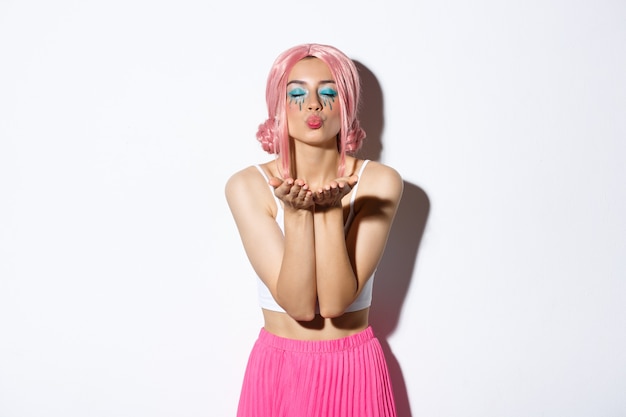 Фото Портрет стильной женской модели в розовом парике, празднующей праздник, дует воздушный поцелуй с закрытыми глазами, стоя на белом фоне
