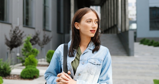 Съемка портрета довольно молодой красивой женщины Кавказа в синей джинсовой куртке с сумкой, стоящей на городской улице и радостно улыбаясь в камеру. Красивая счастливая жизнерадостная женщина на открытом воздухе.