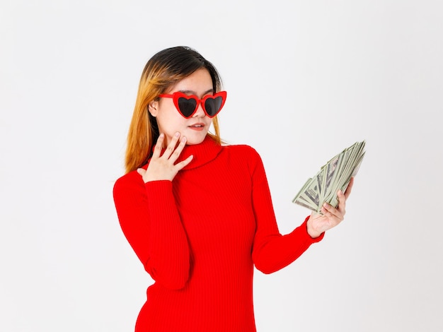 Фото Портрет студия выстрел азиатской уверенно женской модели бизнес-леди в повседневном модном модном красном платье с солнцезащитными очками в форме сердца, держащими долларовую денежную банкноту на белом фоне.