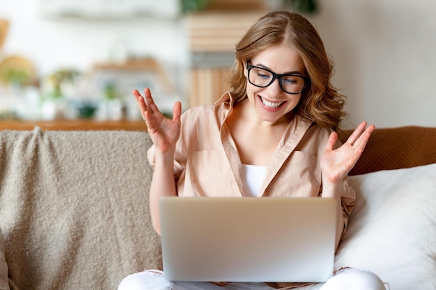 Фото Позитивная молодая женщина в повседневной одежде и очках сидит дома на диване и разговаривает в видеочате через нетбук, жестикулируя и улыбаясь