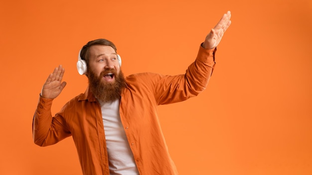 Фото Позитивный бородатый парень в беспроводных наушниках танцует над оранжевой стеной