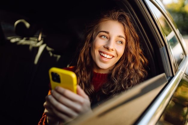 Позитивная молодая женщина в повседневной одежде сидит на заднем сиденье машины с мобильным телефоном