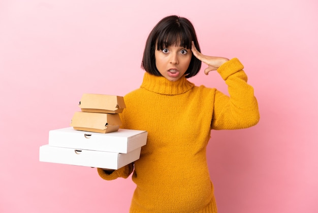 Photo pregnant woman holding pizzas and burgers isolated on pink background with surprise expression