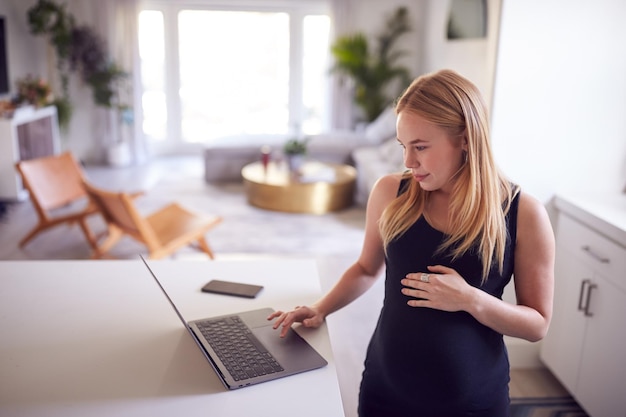 Беременная женщина, стоящая у кухонного стола и работающая из дома на ноутбуке