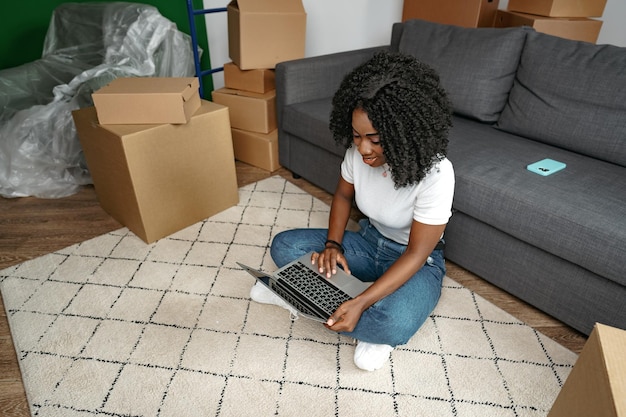 リビングルームのラップトップで床に座っている新しい家にある箱を持ったアフリカの女性