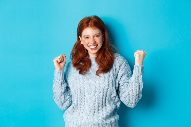 Фото Удовлетворенная рыжая девушка достигает цели и празднует, делая жест кулака и радостно улыбаясь, торжествуя победу, стоя на синем фоне.