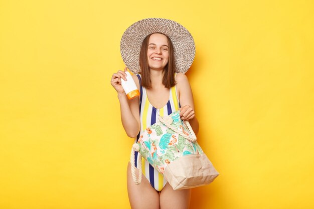 Photo satisfied woman wearing swimsuit isolated on yellow background standing with beach bag in straw hat holding her protective spf cream enjoying of quality