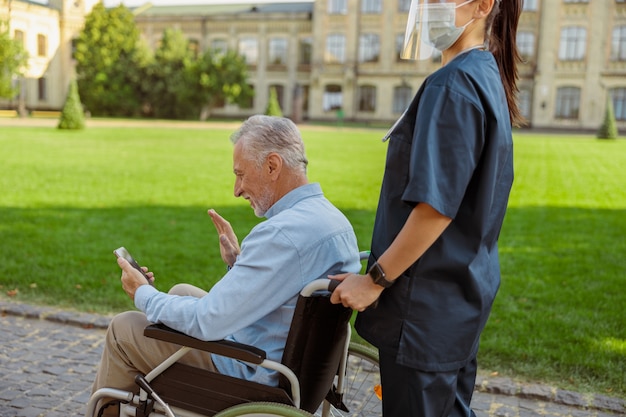 Фото Старший мужчина выздоравливает пациента в инвалидной коляске, делая видеозвонок с помощью смартфона во время прогулки с